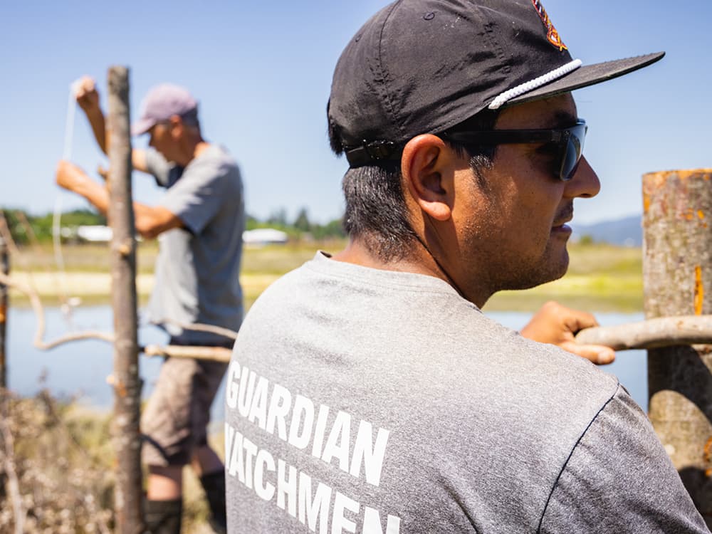Two men in baseball caps building a fence out of reclaimed logs and sticks. The man in front is wearing a shirt that reads "Guardian Watchmen."